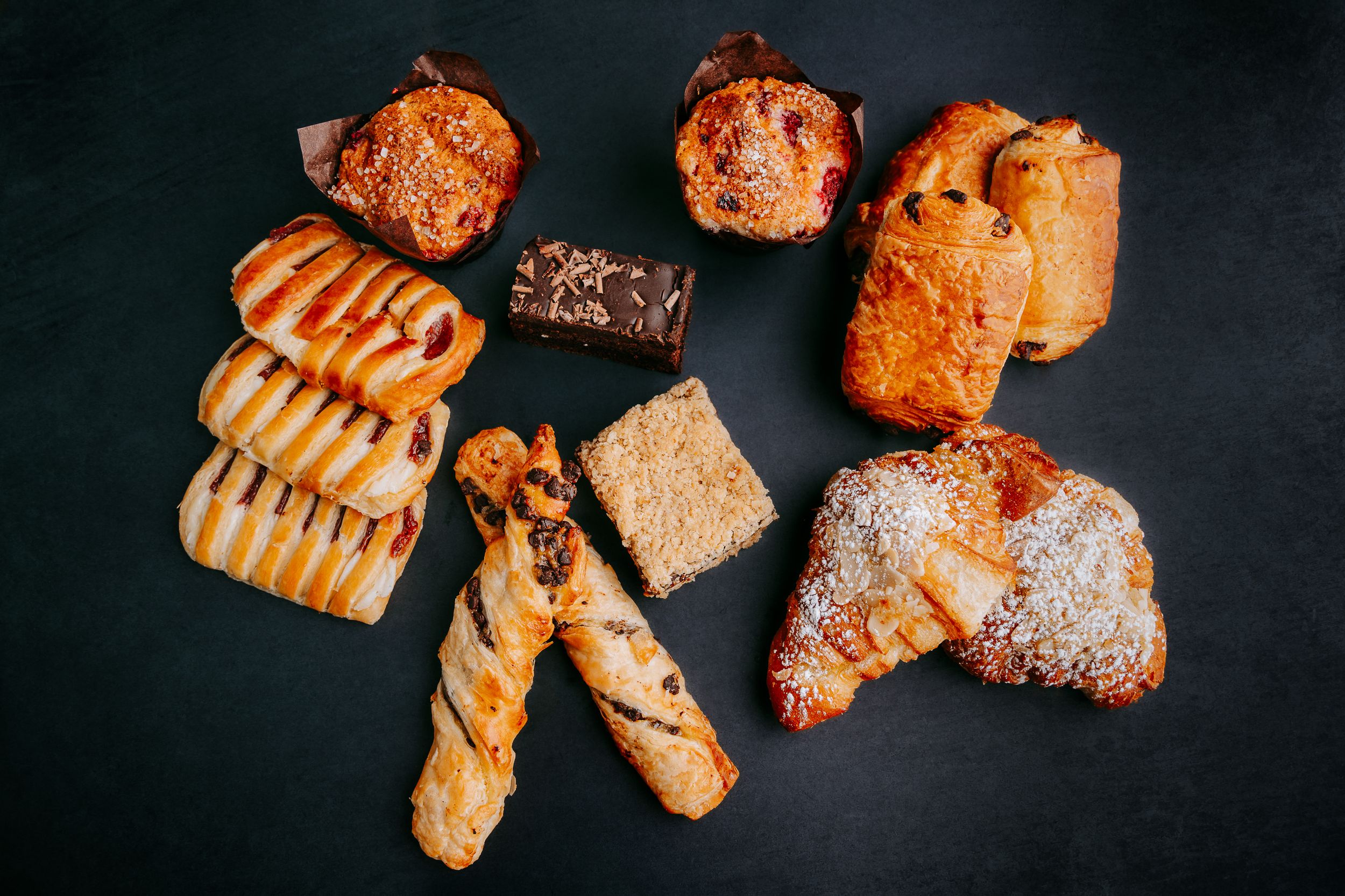 Assorted pastries and baked goods including croissants, danishes, and sweet breads on dark background.