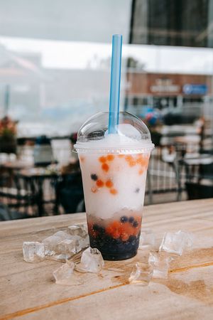 A bubble tea drink with black boba pearls sits on a wooden table with scattered ice cubes beside it.