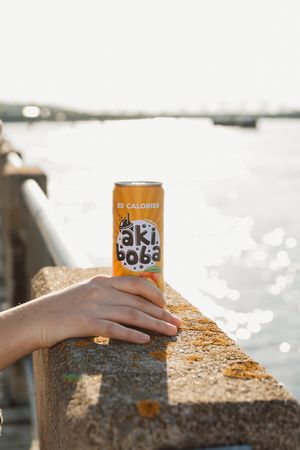 Orange Oki Poki cans resting on a stone wall with sparkling water in the background.