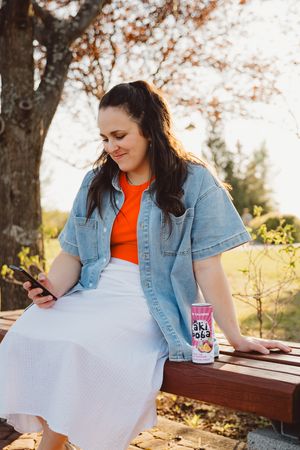 Person in denim jacket relaxing outdoors with Oki Poki drink and mobile phone.