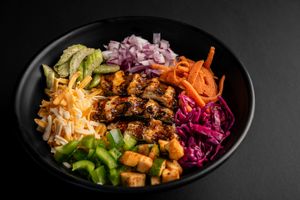 Grilled teriyaki chicken bowl with colorful vegetables and purple cabbage on black plate.