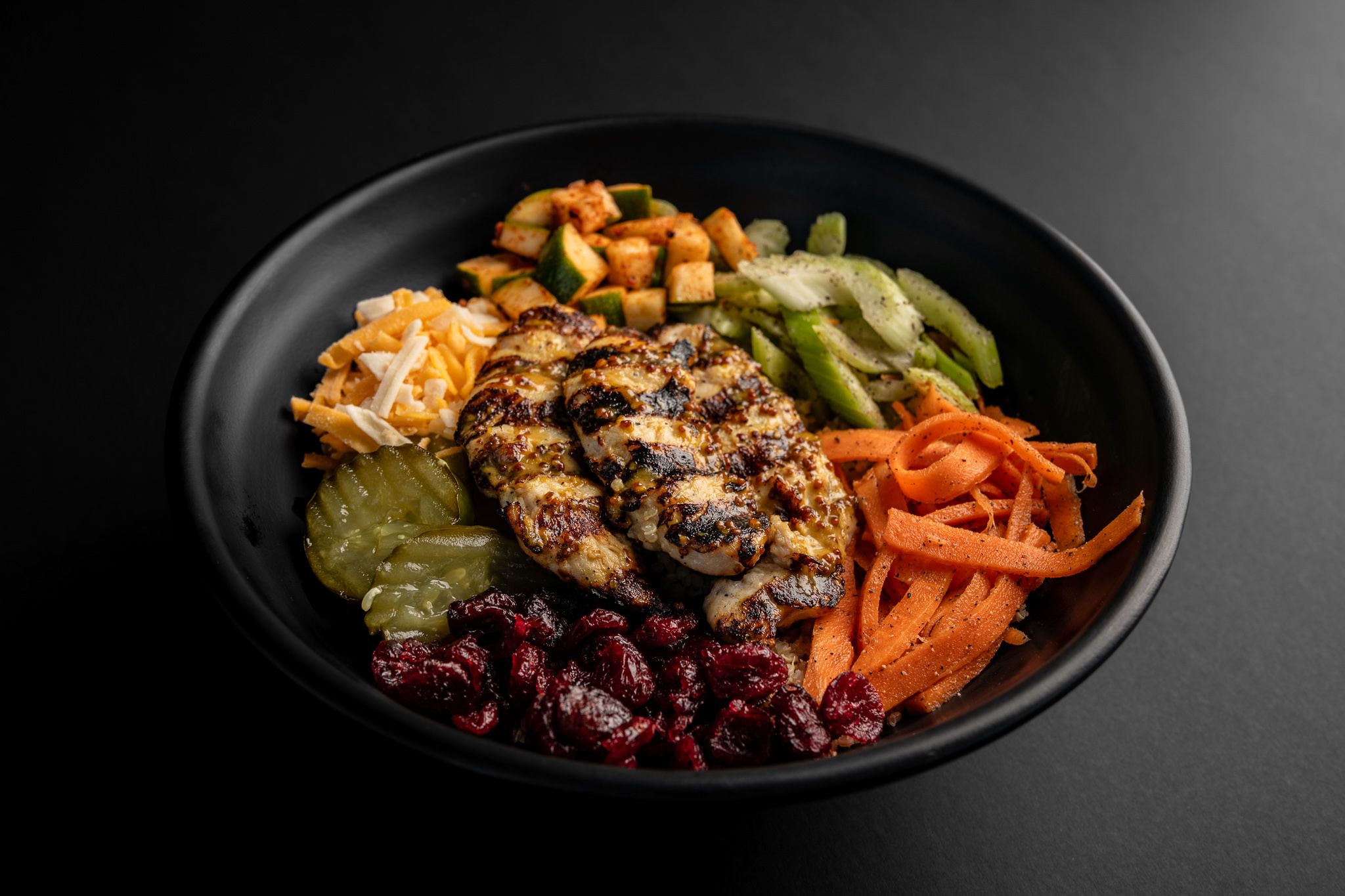 Grilled meat bowl with pickled vegetables, carrots, and cucumber slices on black plate.