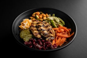 Grilled meat bowl with pickled vegetables, carrots, and cucumber slices on black plate.