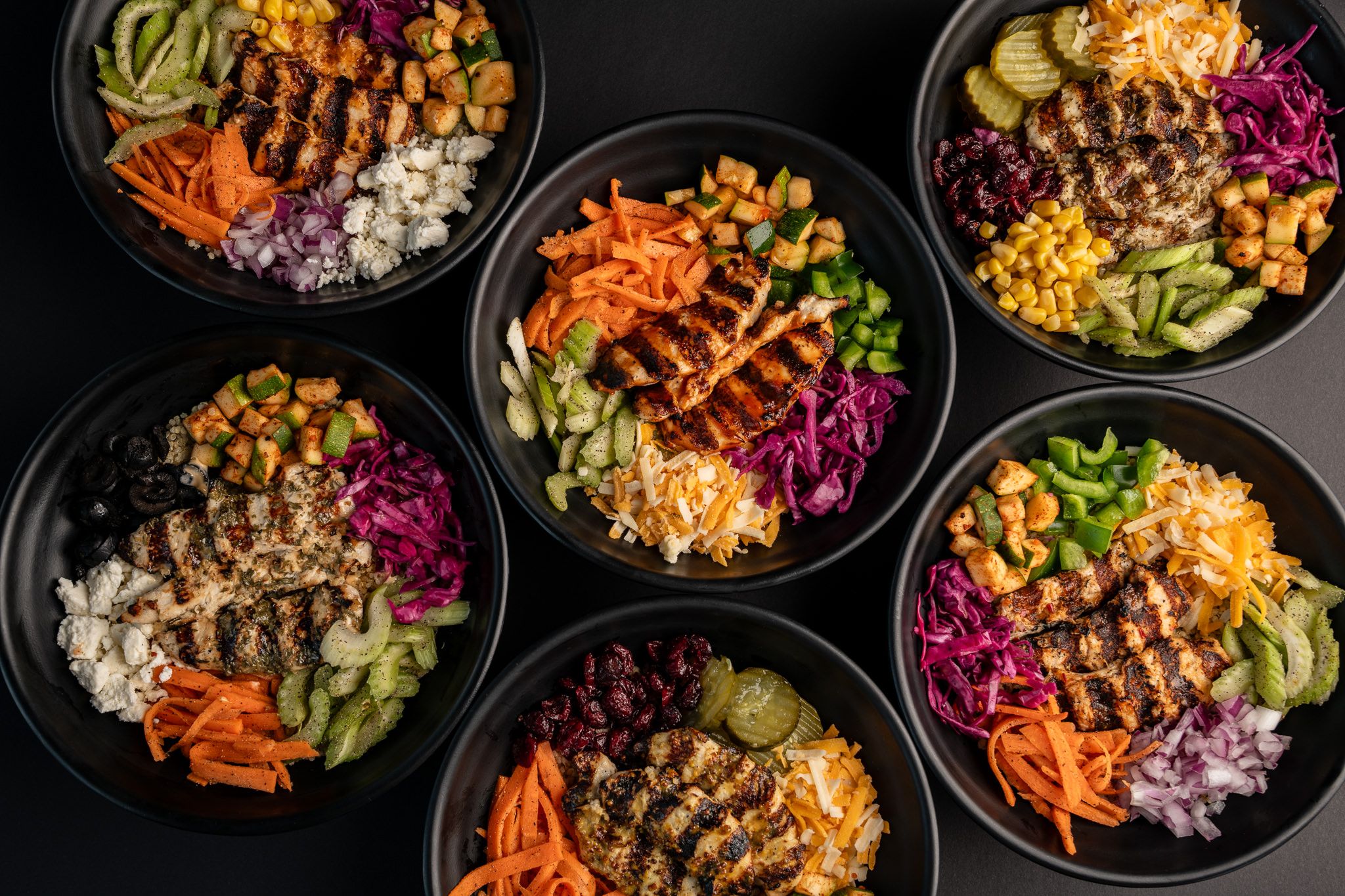 Overhead view of multiple rice bowls with grilled meat and colorful vegetables.