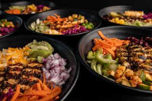 Multiple colorful rice bowls with grilled meat and fresh vegetables arranged on dark surface.