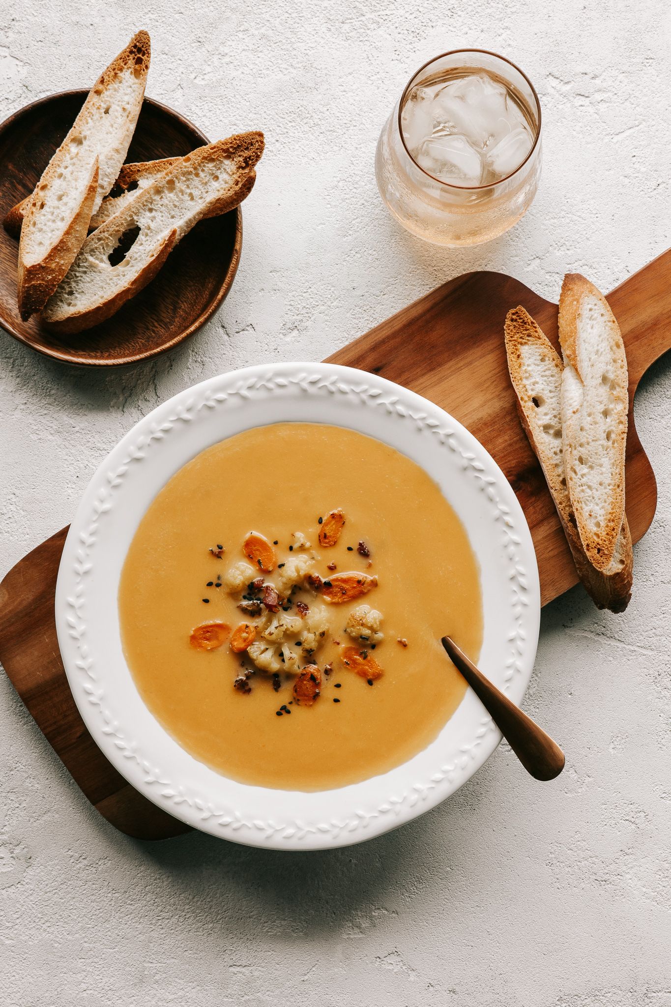 Creamy butternut squash soup in white bowl with toasted baguette slices on wooden board and iced drink.