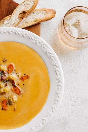 Creamy butternut squash soup in white bowl with roasted vegetables, served with crusty bread slices and iced drink.