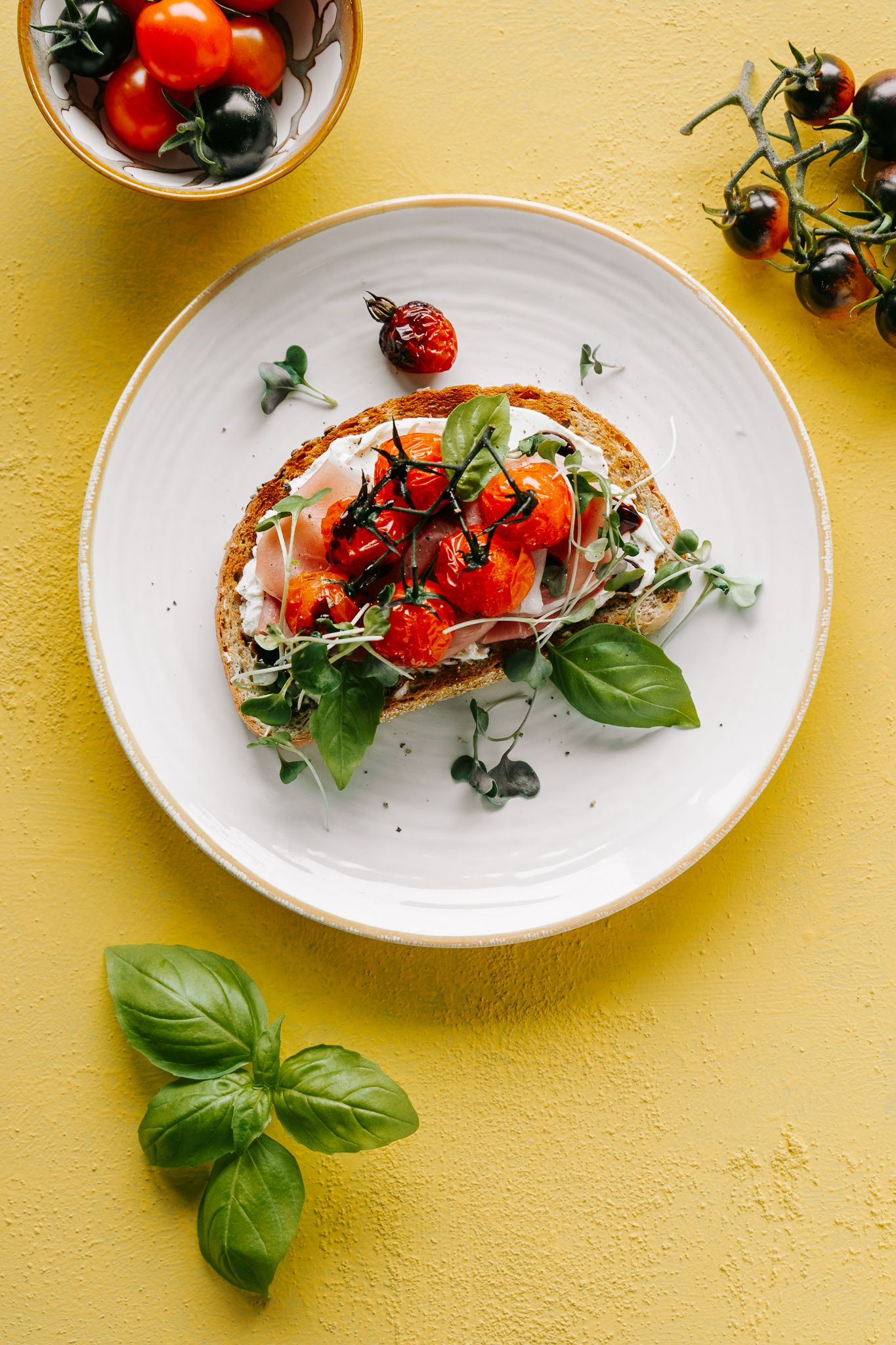 Bruschetta with roasted cherry tomatoes, fresh basil, and microgreens on white plate, yellow background.