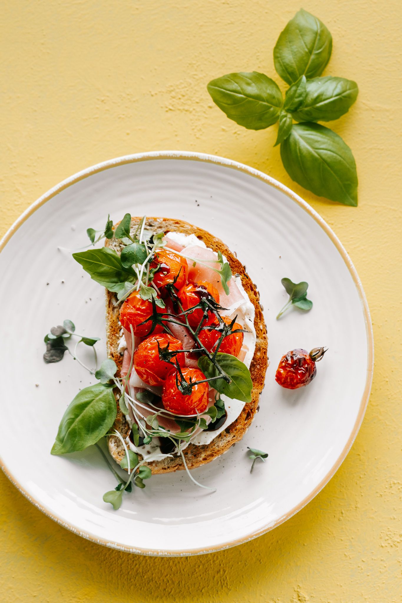 Toasted bread with cream cheese, roasted cherry tomatoes, fresh basil, and microgreens on a white plate.