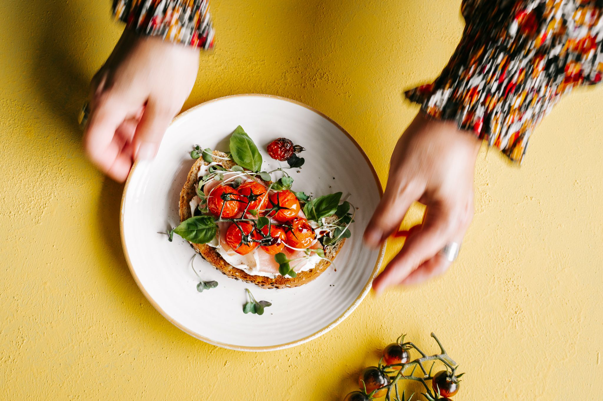 Hands holding a white plate with bruschetta topped with tomatoes and fresh herbs on a yellow background.