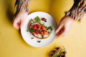 Hands placing a white plate with bruschetta topped with roasted tomatoes and fresh basil on a yellow surface.
