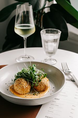 Elegant plated sesame-crusted croquettes with microgreens on sauce, served with white wine and water glasses.