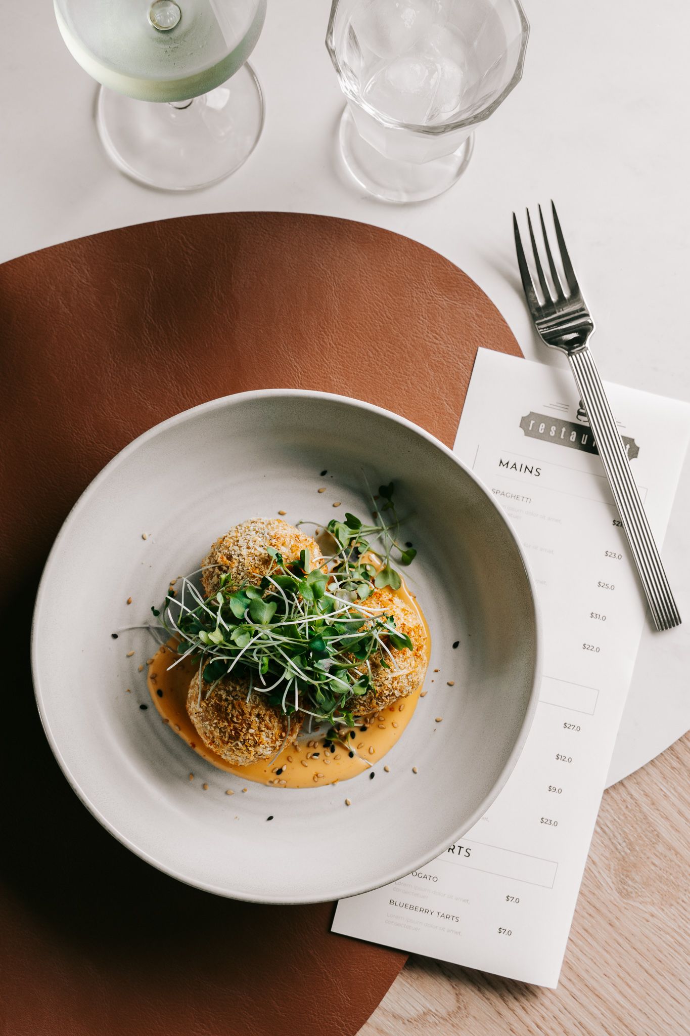 Elegant restaurant dish with microgreens on orange sauce in white bowl, beside menu card, fork, and wine glasses.