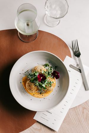 Elegant restaurant dish with microgreens and edible flowers on a white plate, served with white wine on a leather placemat.