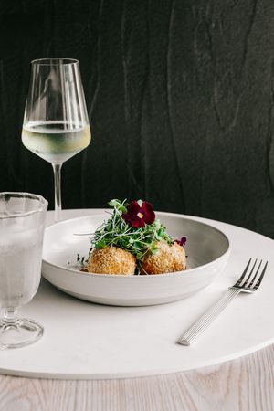 Elegant restaurant dish of breaded arancini with microgreens and edible flower, served with white wine on white plate.