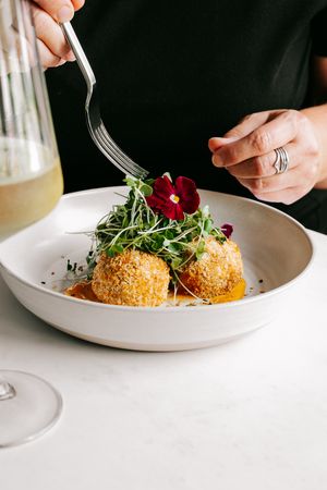 Hands using a fork to eat breaded arancini topped with microgreens and edible flowers in a white bowl.