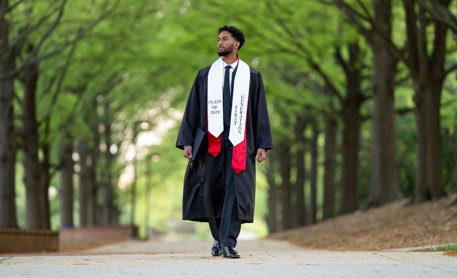 Graduate in red and white regalia poses on campus steps with classic columns and greenery in background.