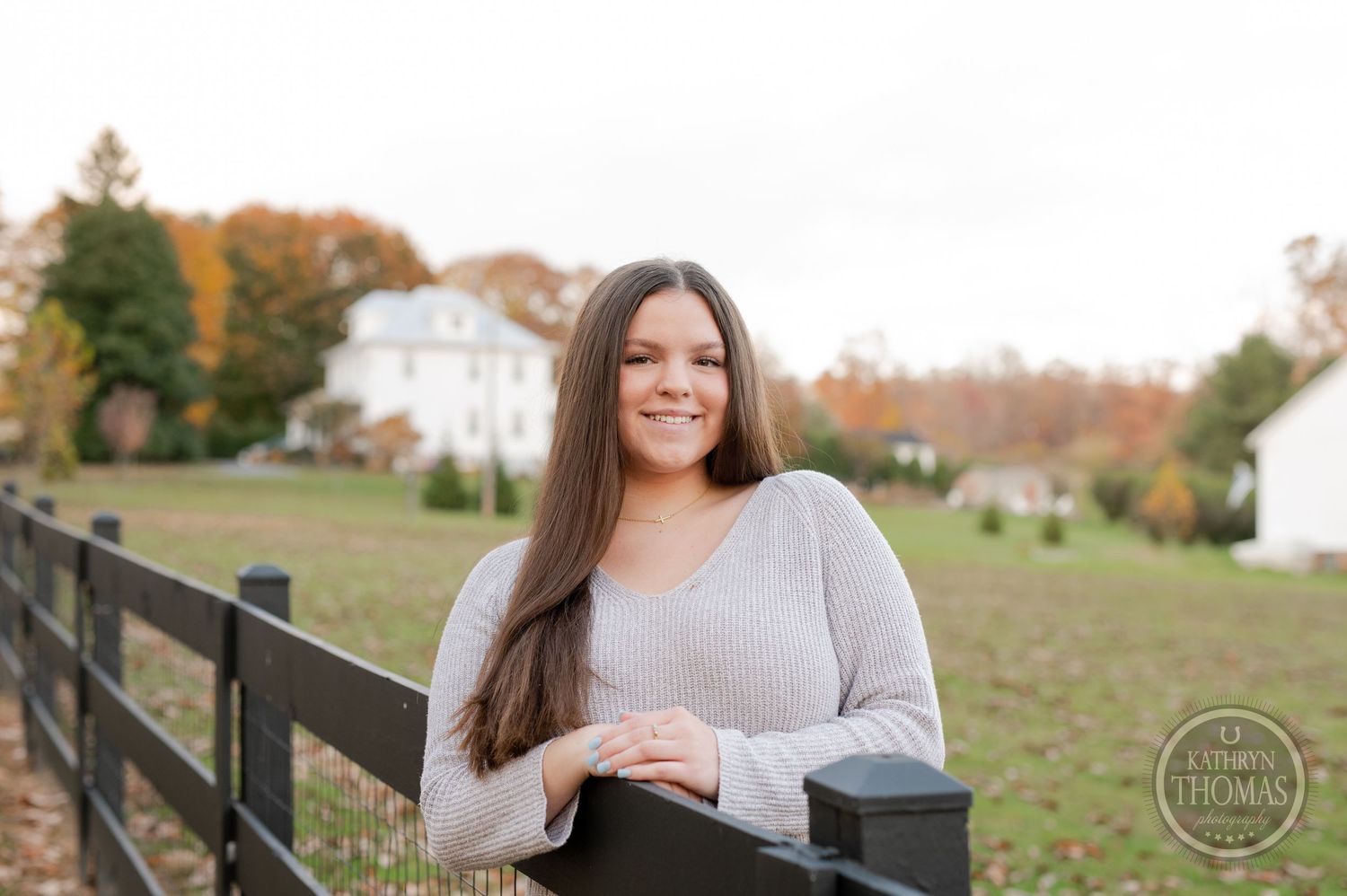 Outdoor portrait by rustic fence with white farmhouse and autumn landscape in background.