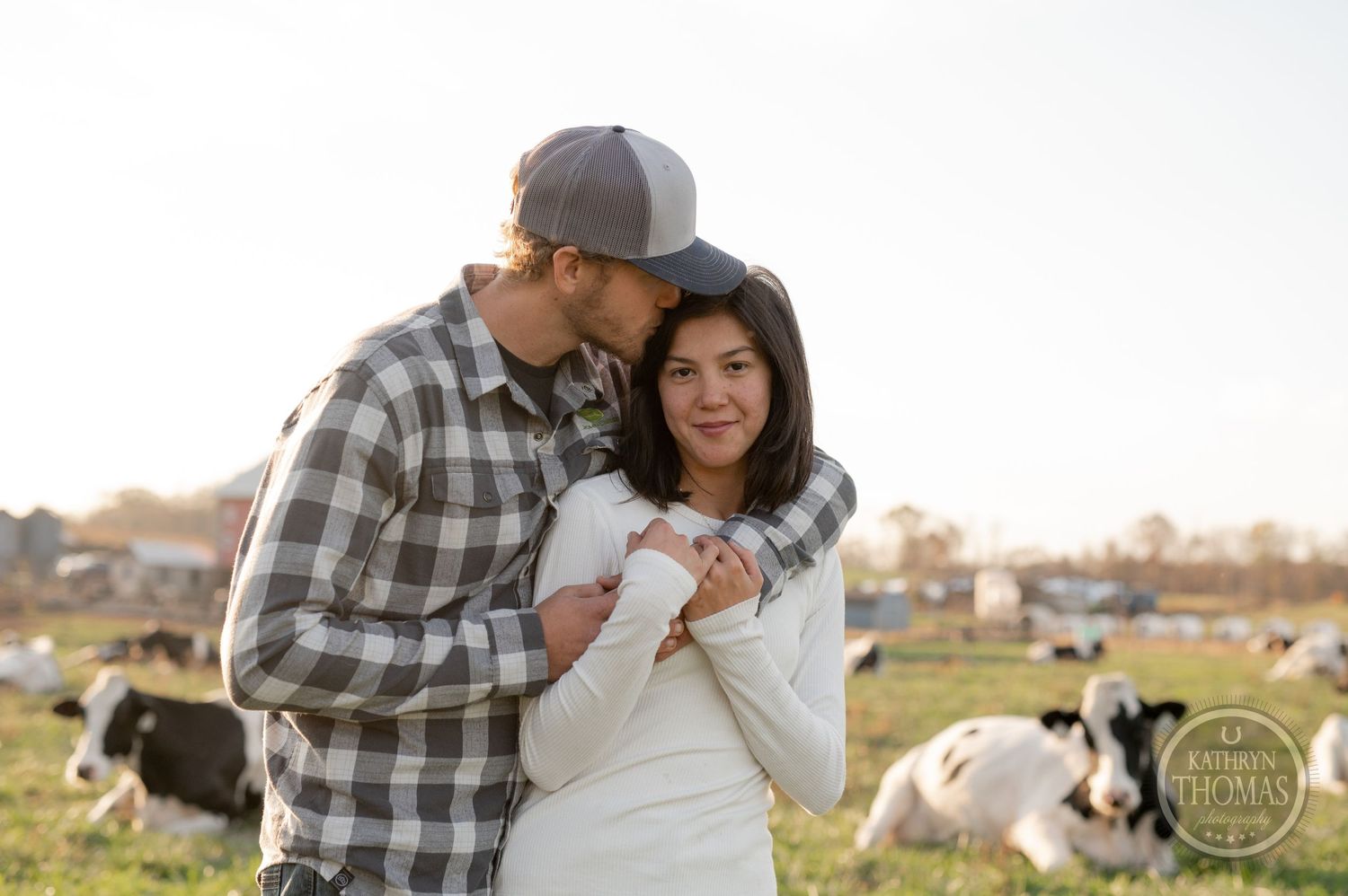 A romantic couple shares a tender moment on their farm with dairy cows grazing in the distance.