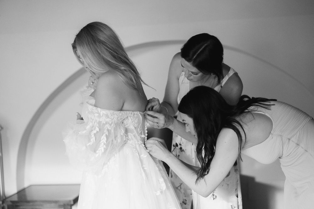 bride getting ready with her two bridesmaids. The bridesmaids are zipping the Bride's dress