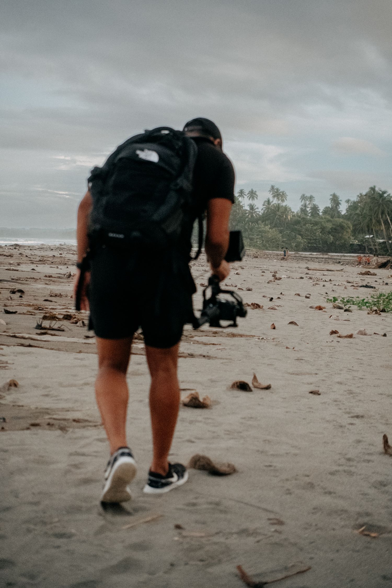Photographer walking on a sandy beach with camera gear under cloudy skies.