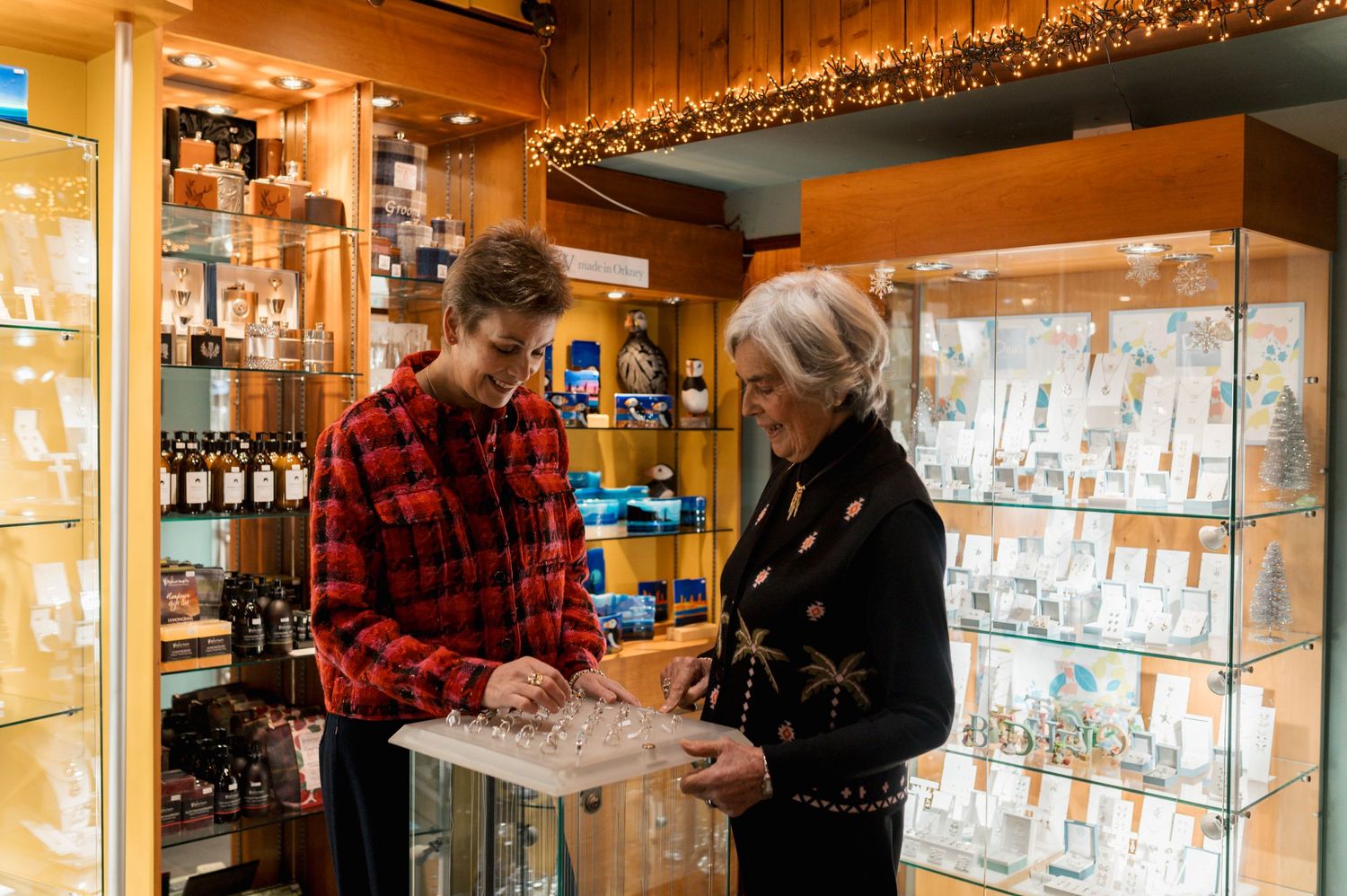 Employee assists customer looking at jewelry display case in cozy store with warm lighting and festive decorations.