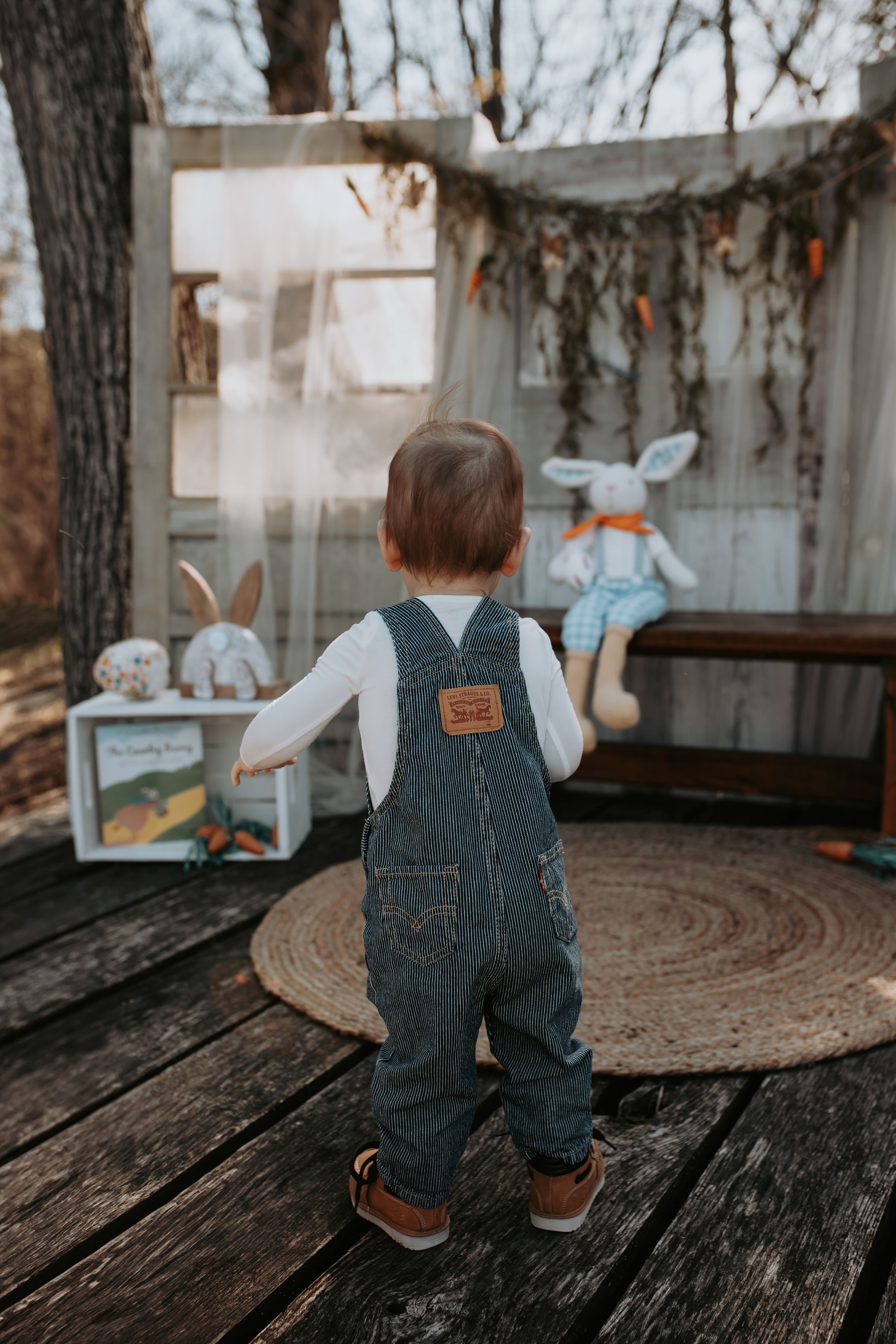 Small toddler in denim overalls sitting on wooden deck with rustic backdrop.