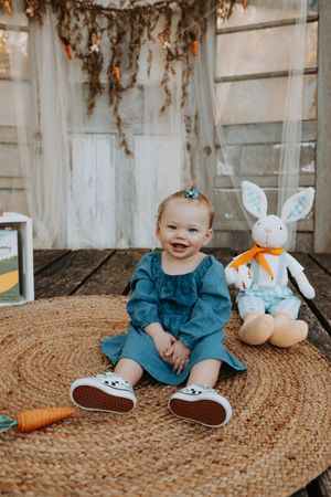 Baby in blue outfit sits with plush bunny and carrot toy on round jute rug.