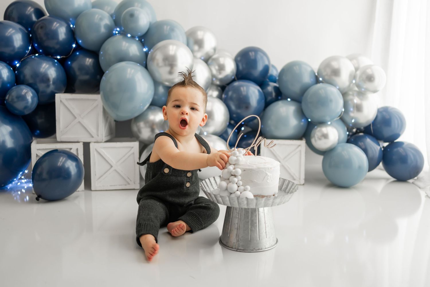 Baby milestone photography setup with blue and silver balloon garland on white backdrop.