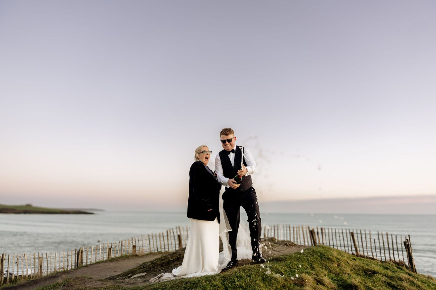A coastal wedding portrait with a couple standing on a cliff overlooking the ocean at sunset.