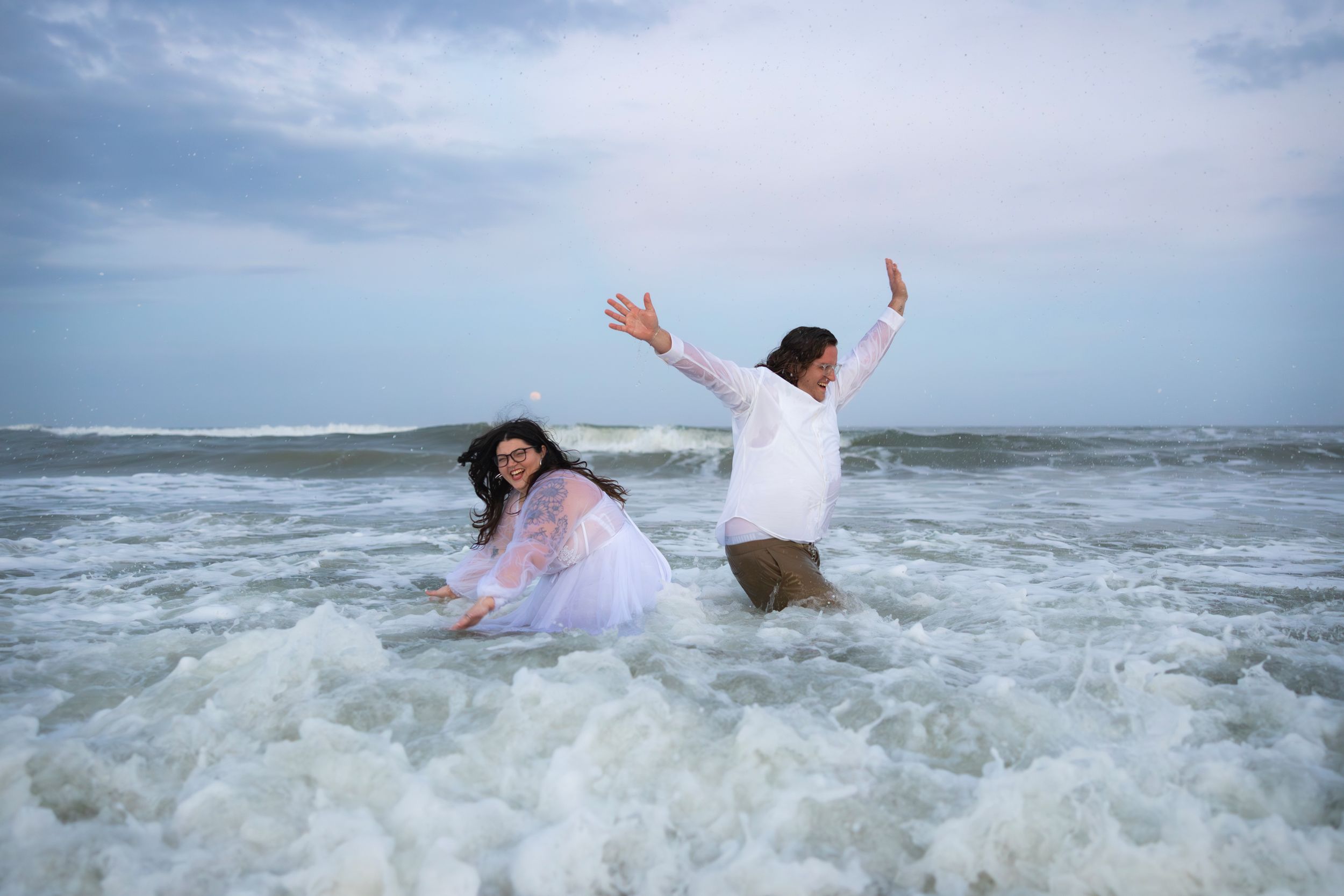 Joyful couple splashing in ocean waves wearing wedding attire.