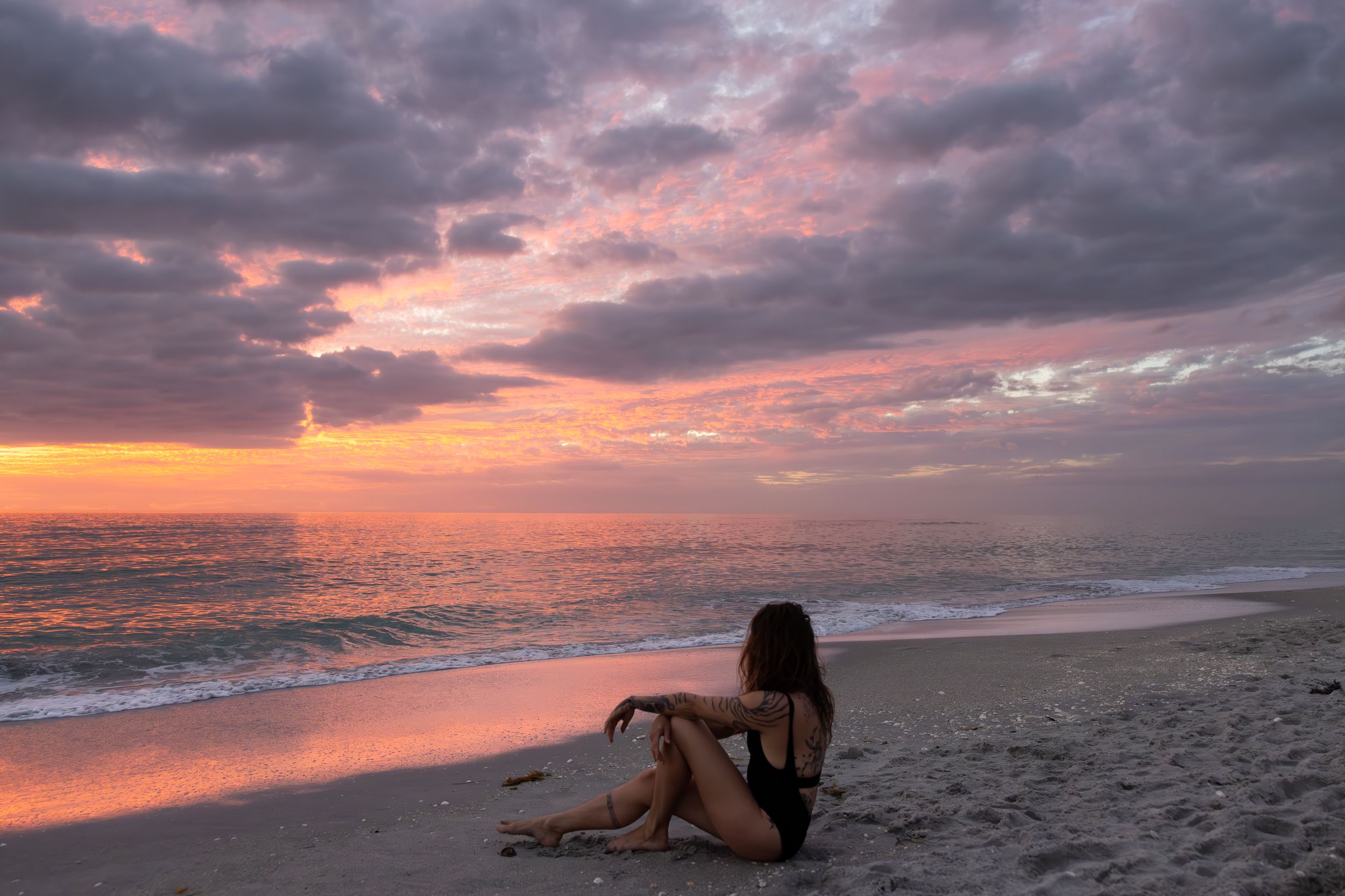 Silhouette sitting on beach during vibrant purple and orange sunset.
