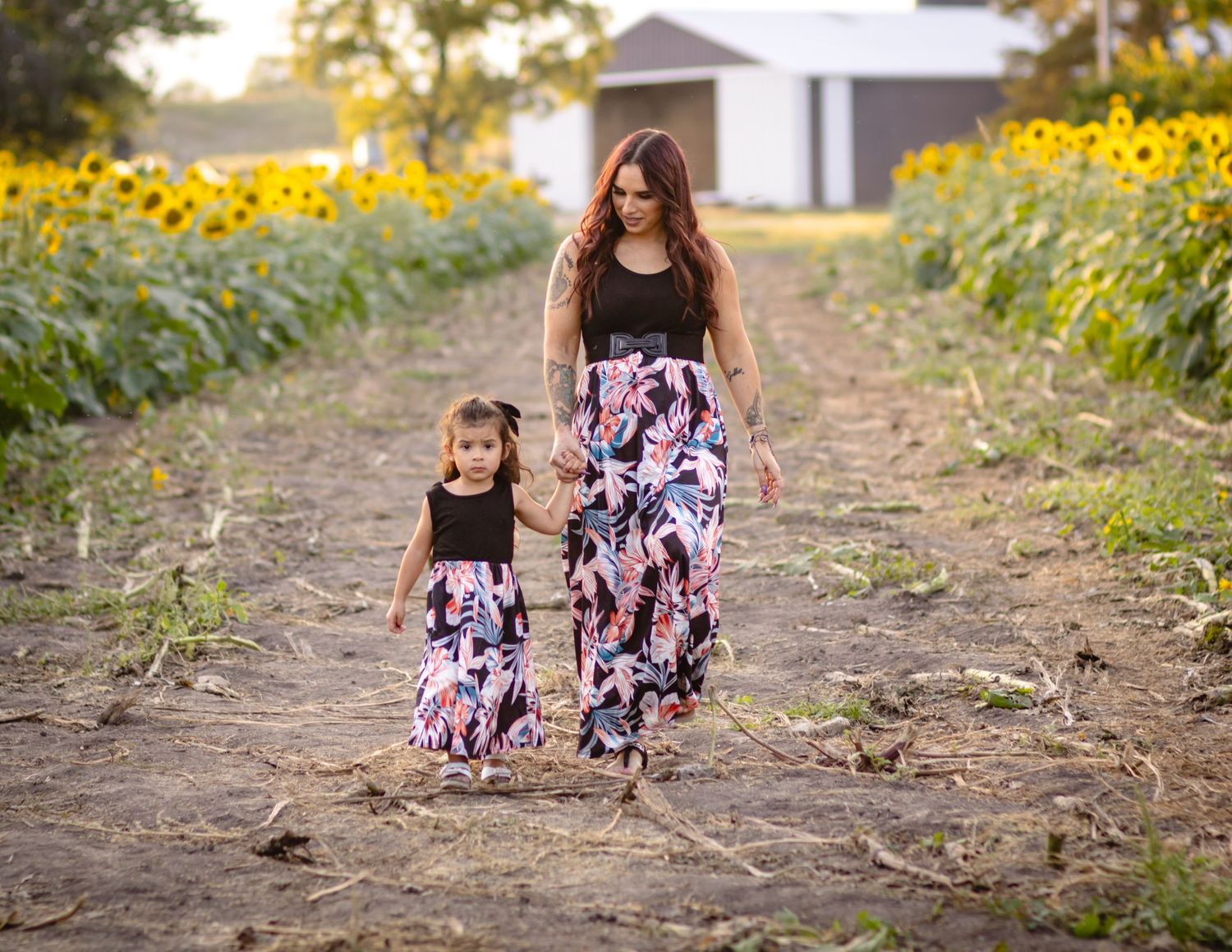 Mother and daughter walk hand in hand down a path surrounded by bright sunflowers wearing matching floral skirts.