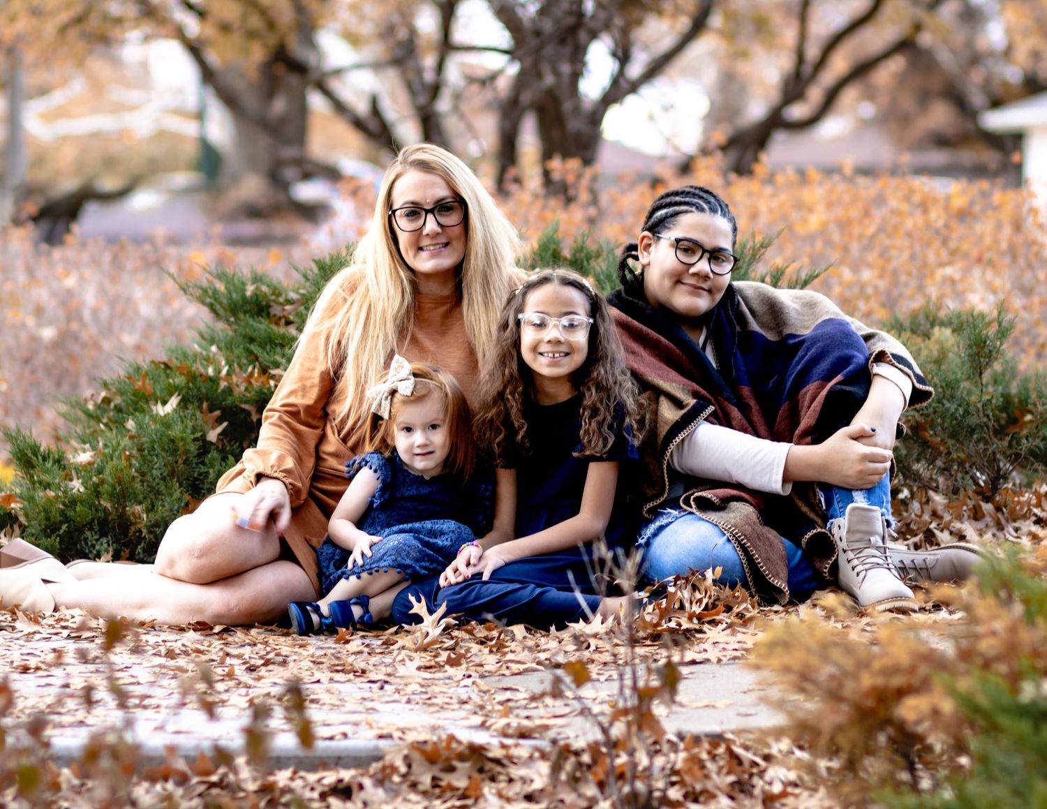 A family of four sits together on autumn ground covered in fallen leaves during an outdoor photo session.