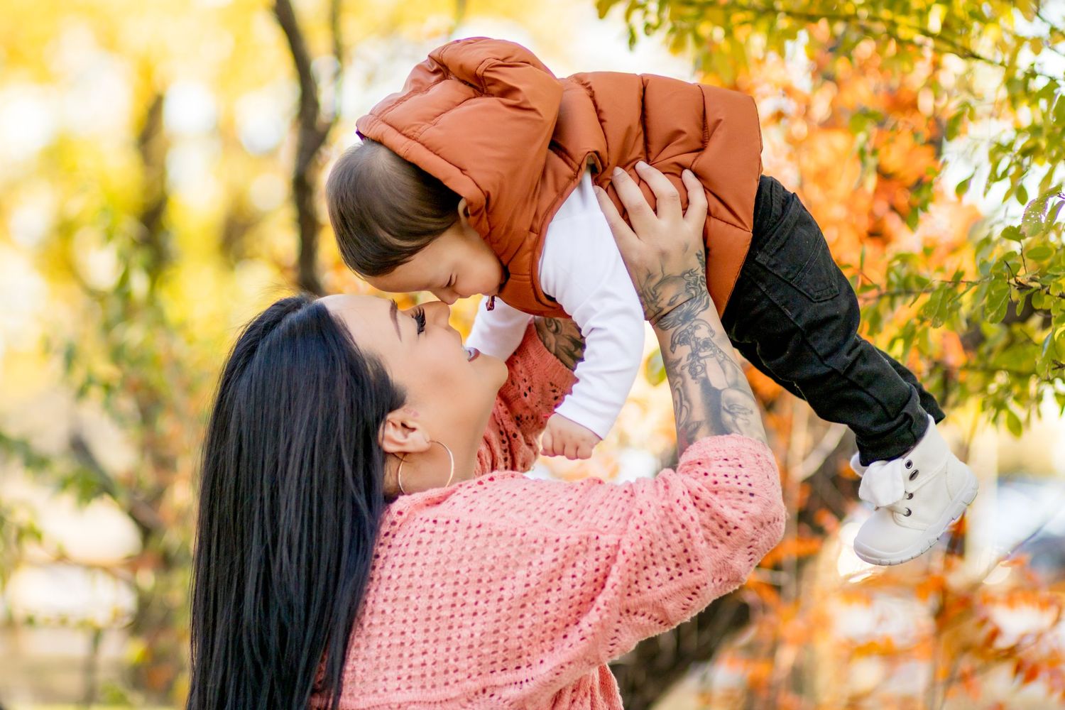 A parent in a pink knit sweater lifts and kisses a toddler during a sunny autumn day with golden leaves in background.