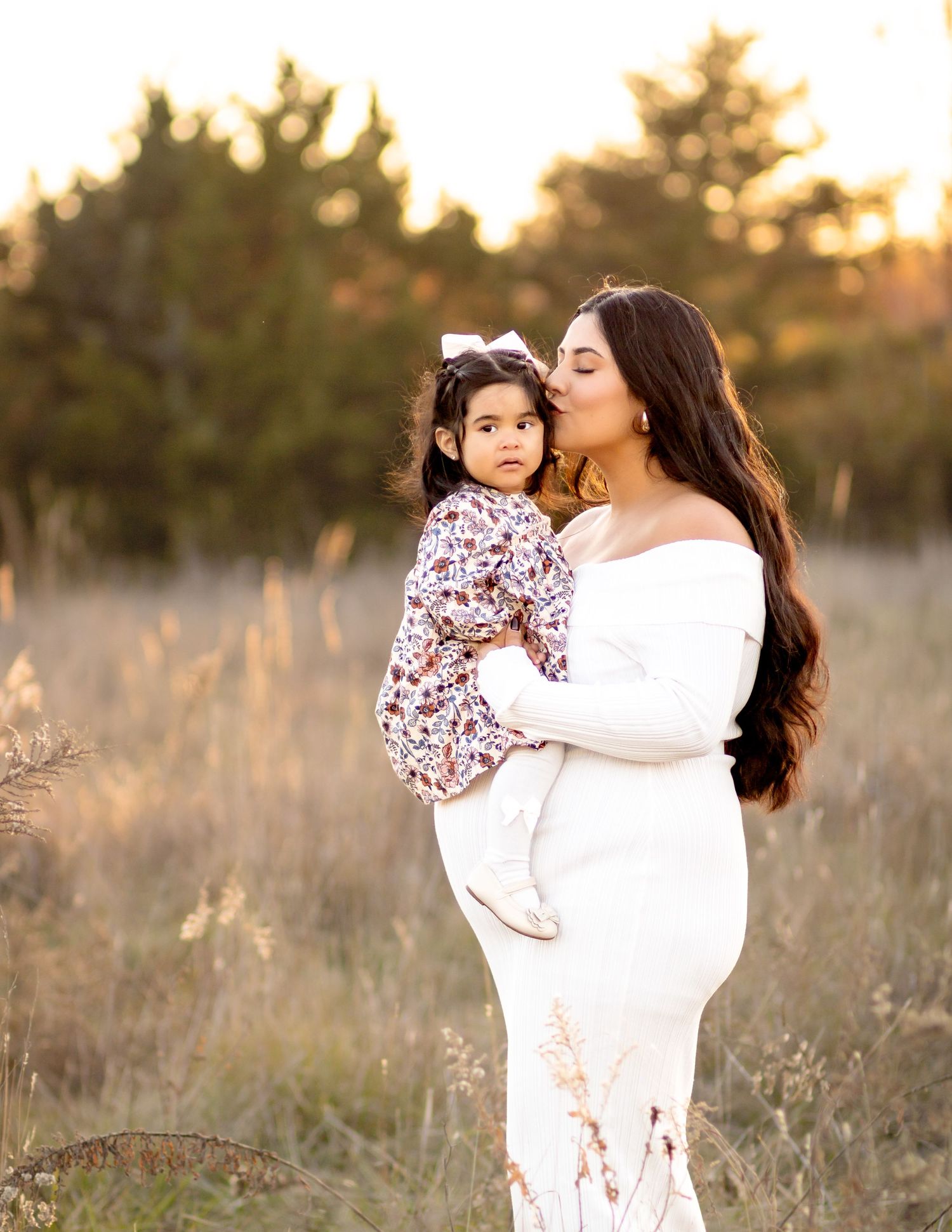 A tender family moment at golden hour with a woman in white dress holding a young child in patterned outfit in a grassy field.
