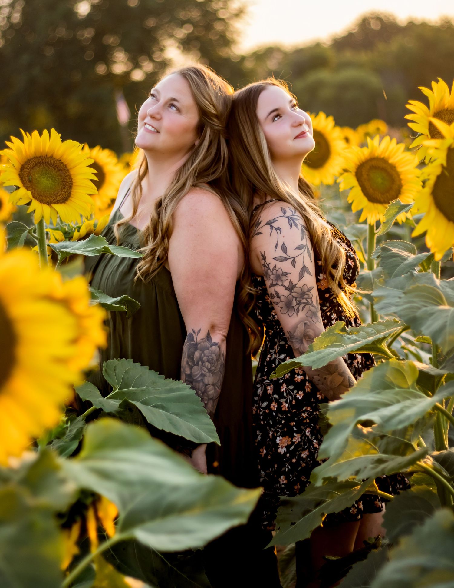 Two friends pose back-to-back in a vibrant sunflower field at golden hour wearing dark dresses with tattoos.