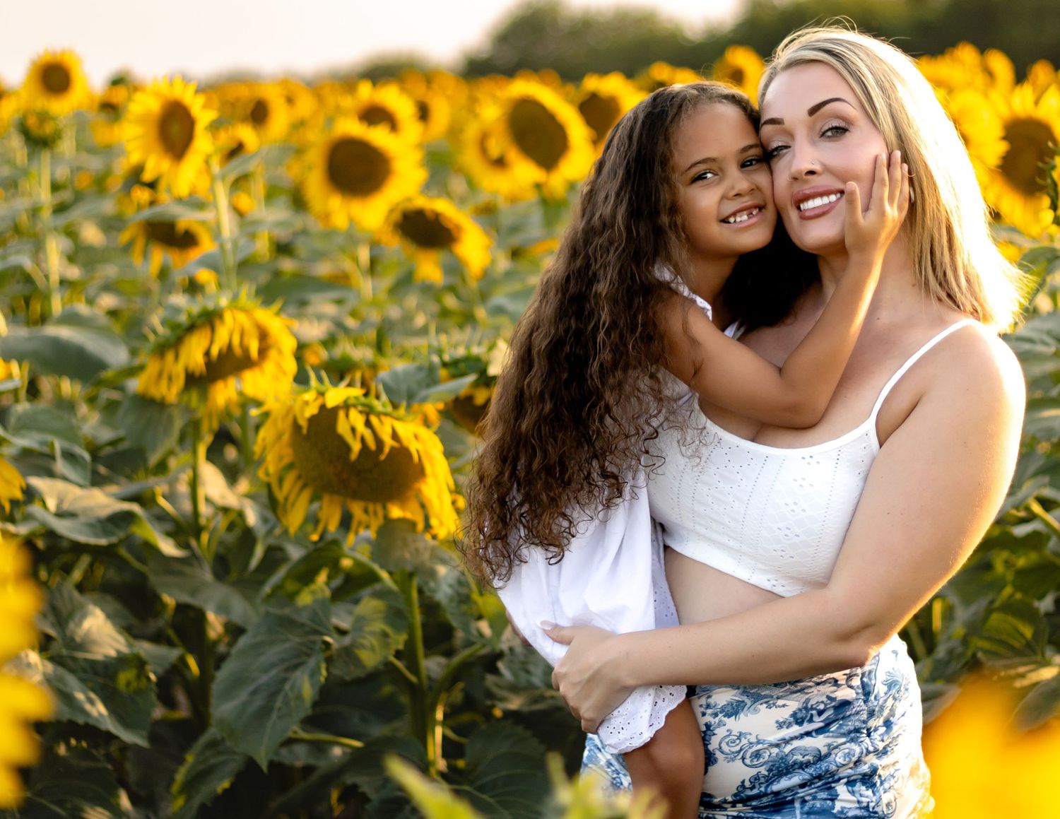 Two friends share a joyful embrace while posing in a vibrant field of blooming sunflowers during golden hour.