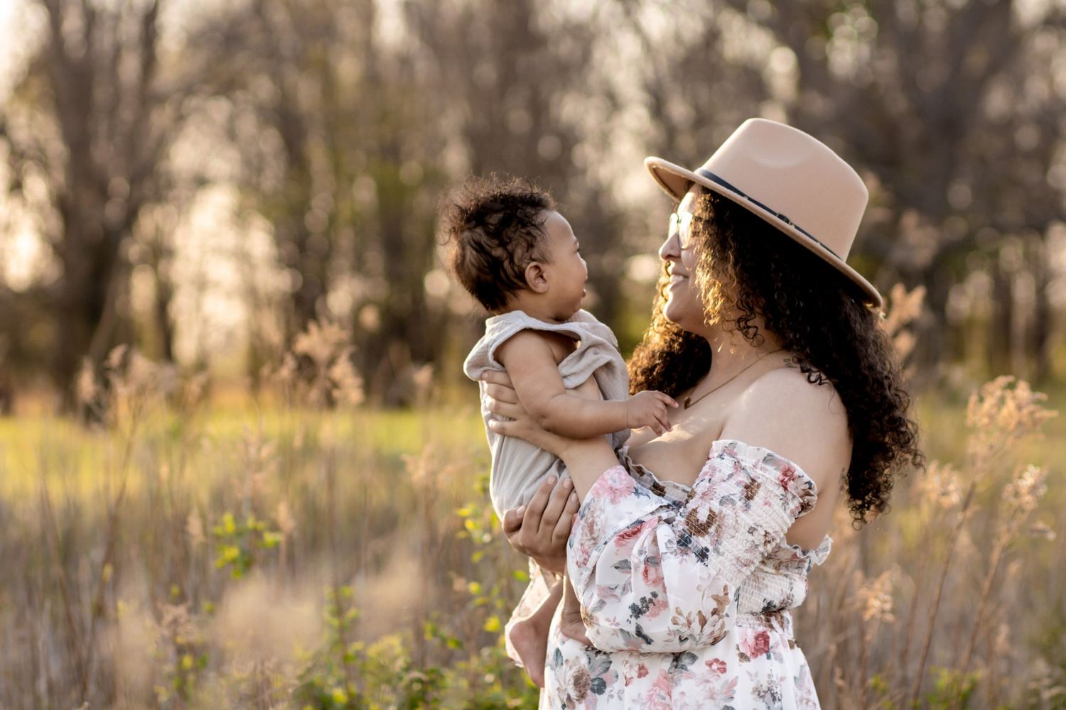 Woman in floral dress and hat holds small child while standing in golden sunlit field at sunset.