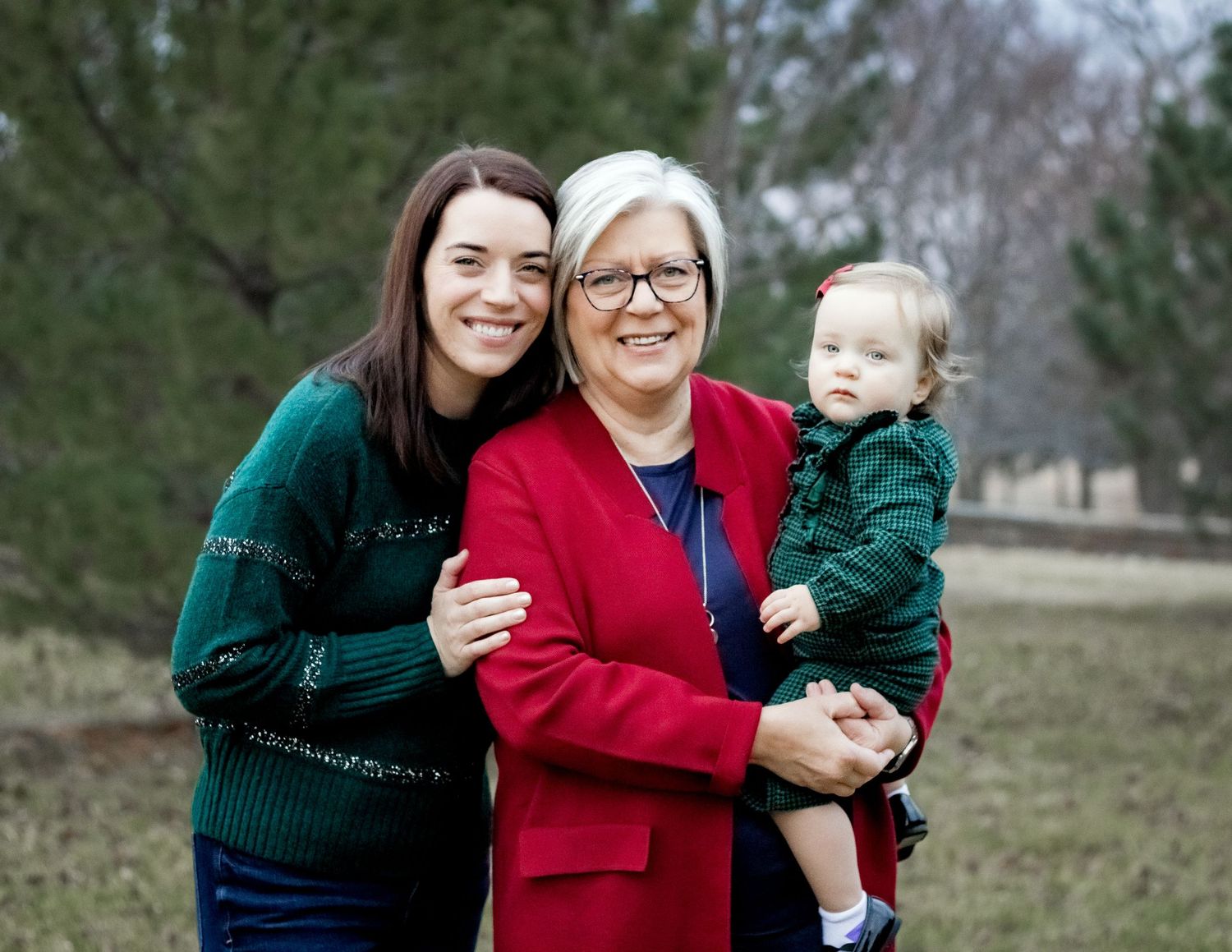 Three generations smile together outdoors, wearing festive winter sweaters in green and red against a natural backdrop.