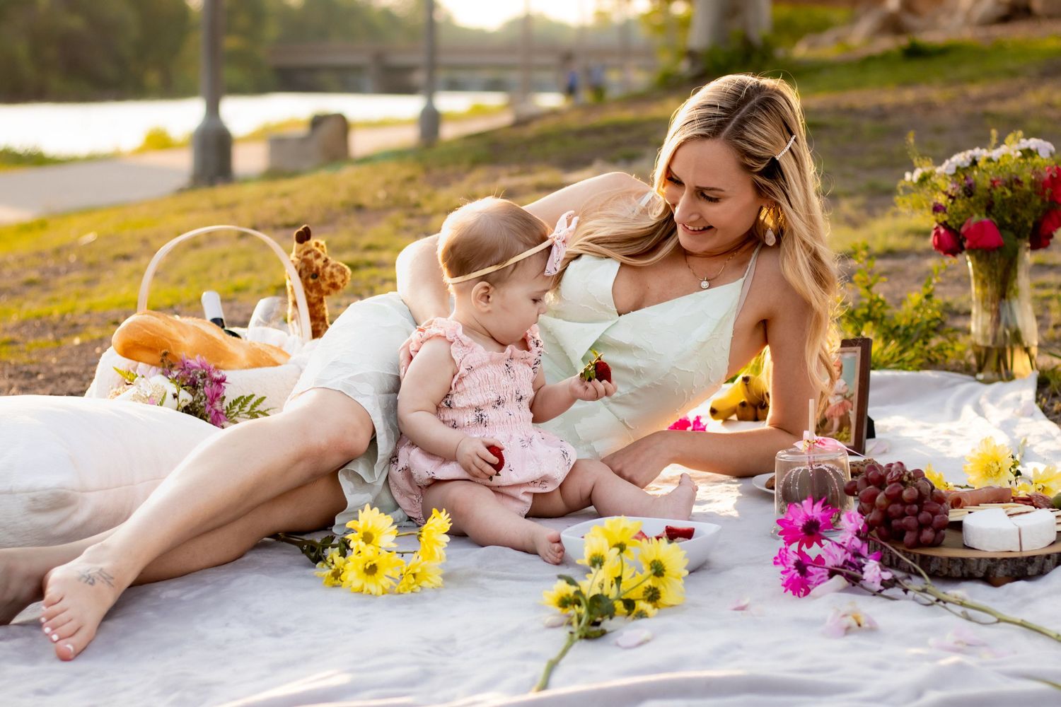 Outdoor picnic scene on a blanket with yellow flowers, food spread, and people relaxing by a lake at sunset.