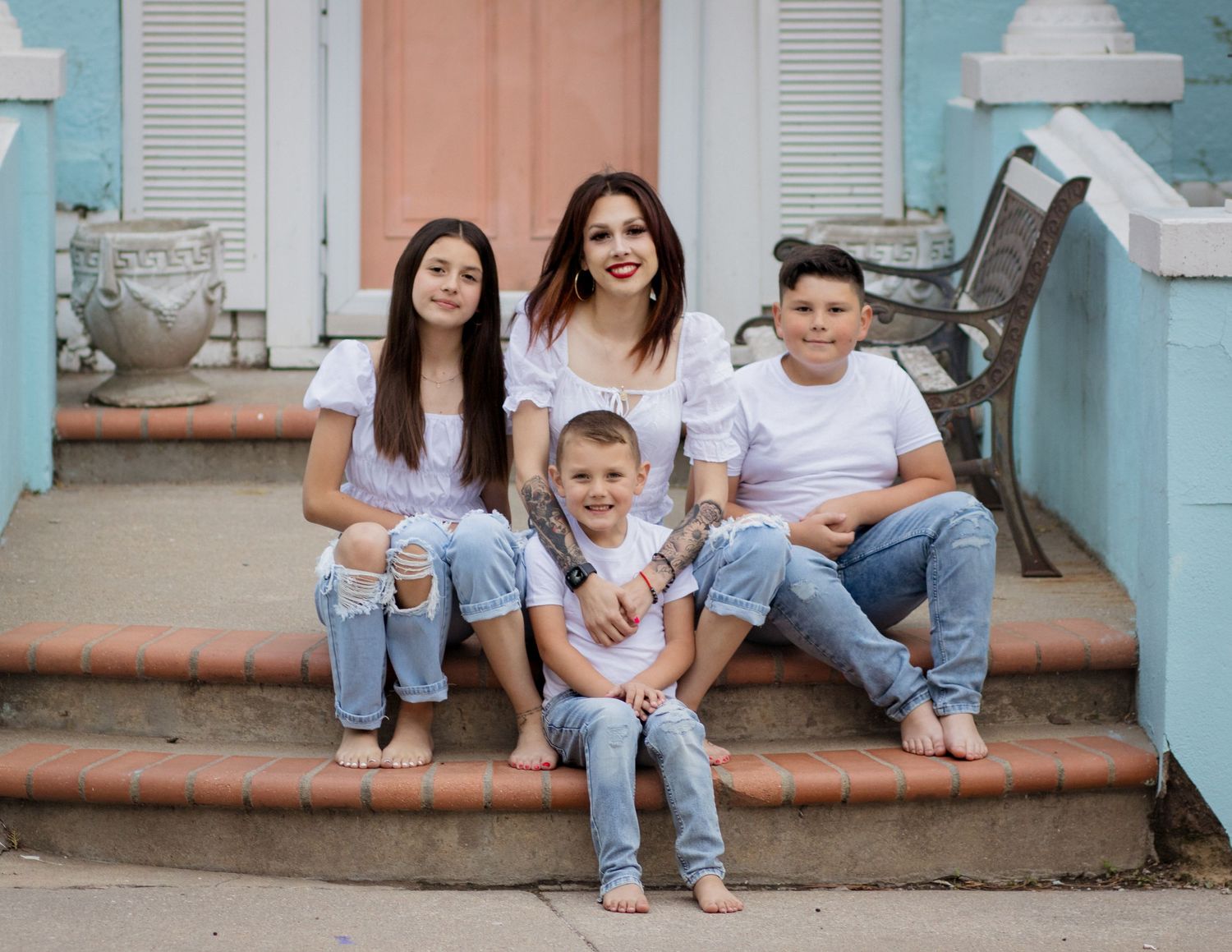 A family sitting together on outdoor steps of a pastel blue and pink building, all wearing casual denim and white outfits.