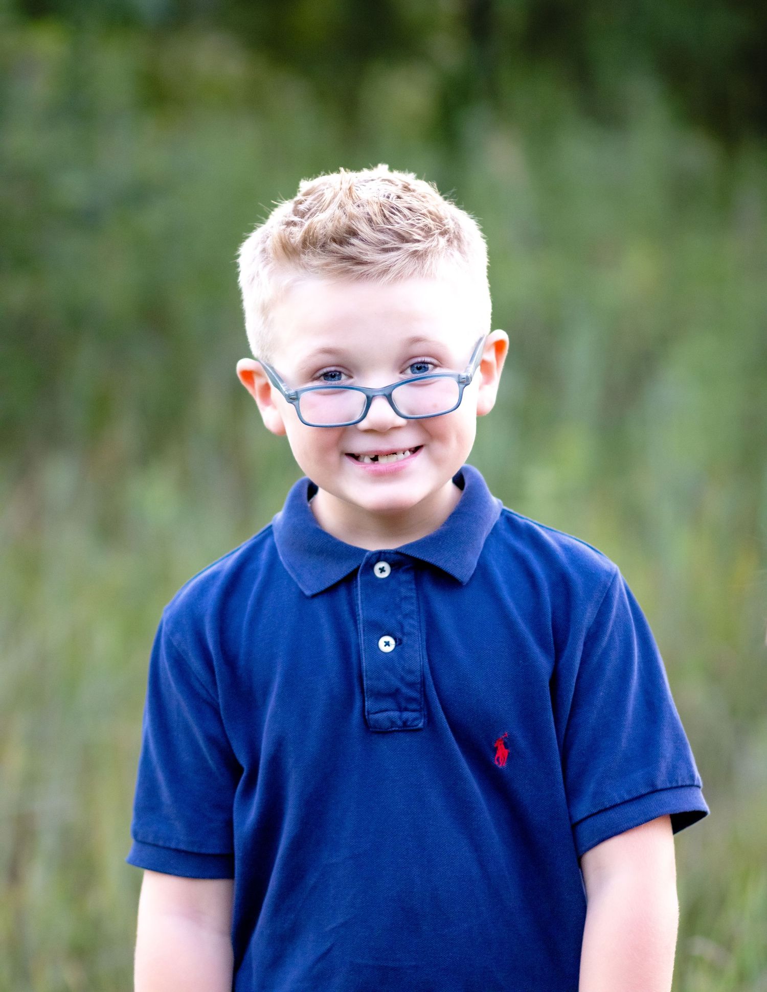 Young person wearing glasses and a navy blue polo shirt with a red logo smiles outdoors against a blurred green background.