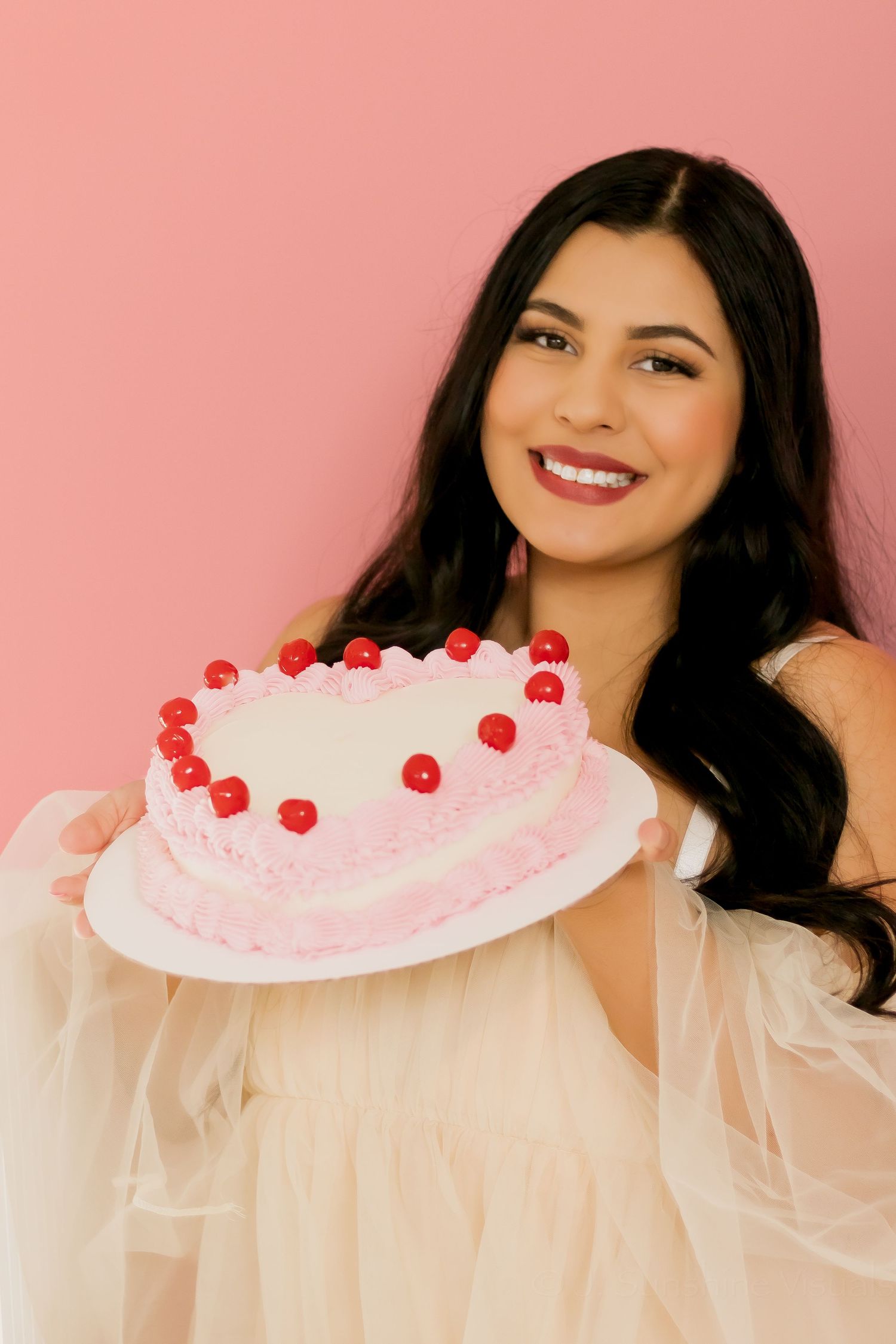 A pink frosted cake adorned with red cherries is held against a coral background in a dreamy, romantic setting.