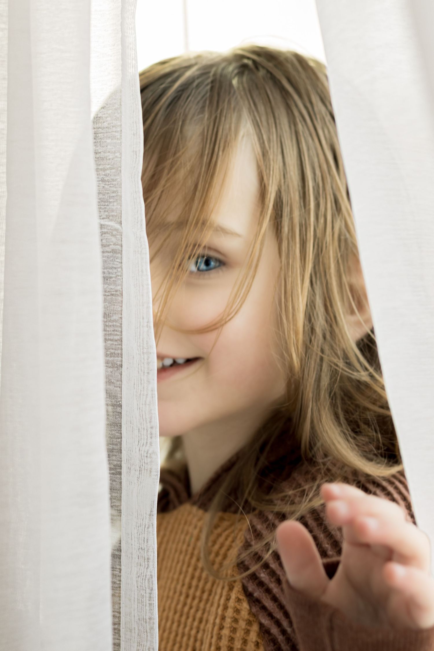 A child with bright blue eyes and blonde hair peeks playfully from behind a white curtain while wearing a brown sweater.