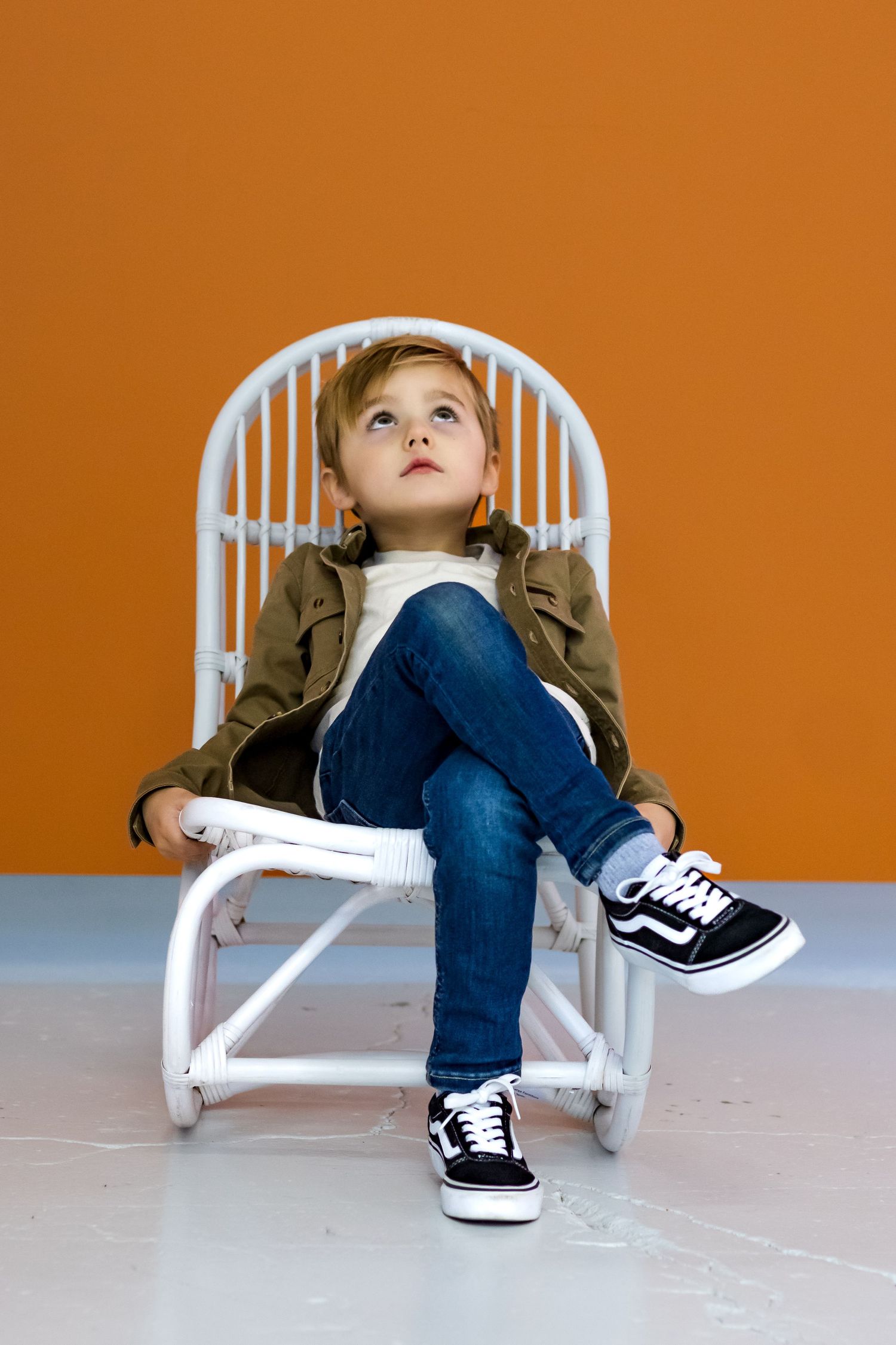 Young child sits thoughtfully in a white wicker chair wearing jeans and black sneakers against an orange wall.