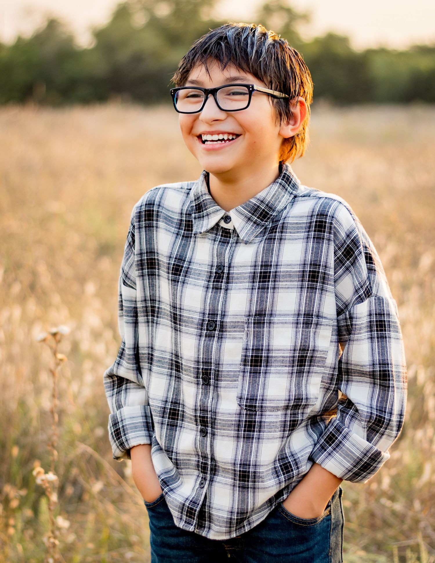 A young student wearing glasses and a plaid flannel shirt smiles joyfully while standing outdoors in a golden field at sunset.