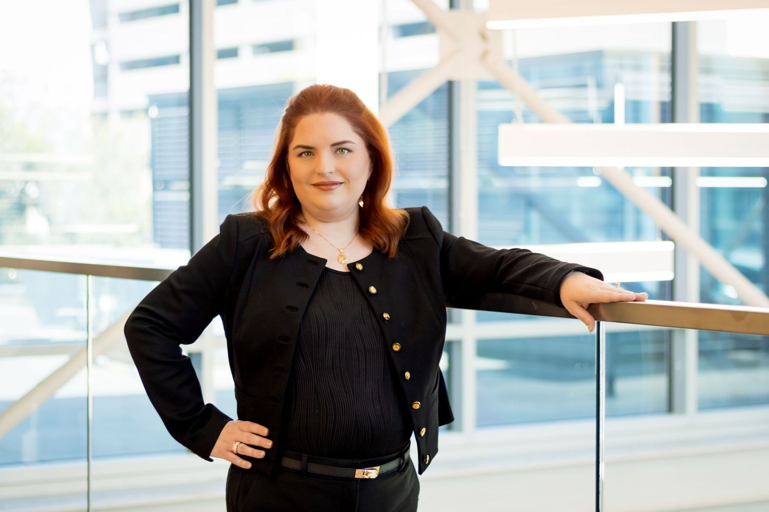 Professional in a black blazer stands confidently in a modern office building with glass windows and handrails.