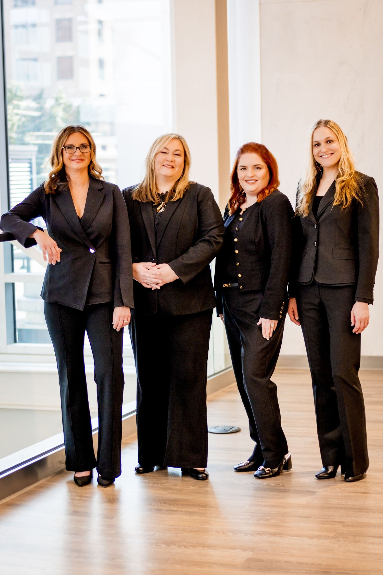 Professional business team in black suits stands confidently in modern office space with hardwood floors and windows.