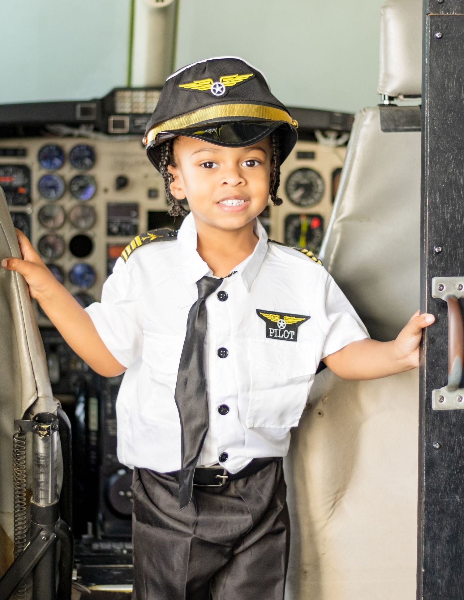 A young child wearing a pilot's uniform and hat poses in an airplane cockpit with aviation instruments visible.
